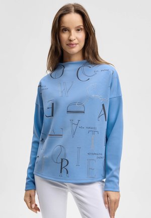 Frau mit langem braunem Haar, die ein hellblaues langärmliges Oberteil mit schwarzen und silbernen Buchstaben sowie weiße Hosen trägt, stehend.