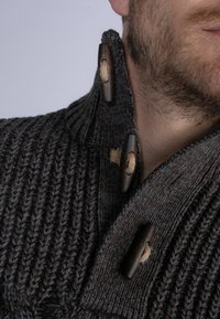 Mies, joka pukeutuu tummanharmaaseen neulepuseroon, jossa on puunäppäimet ja narusilmukat. Kuvassa lähikuva kauluksesta ja osittaisesta parrasta.