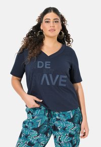 Camiseta de cuello en V de color navy con un gráfico texturizado "RIDE THE WAVE" en la parte frontal, combinada con pantalones de estampado de hojas en tonos verdeazulado y verde.