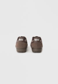 Adidași din suede maro cu o finisare texturată, având o talpă din cauciuc plată și un logo alb "NIKE" pe călcâi. Design simplu și funcțional.