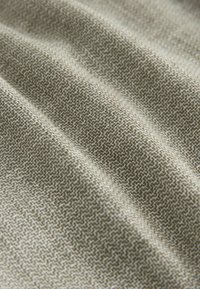 Tekstilstof med et diskret geometrisk mønster, der har en dæmpet grøn og beige farveskala, og viser en blød, tekstureret overflade.