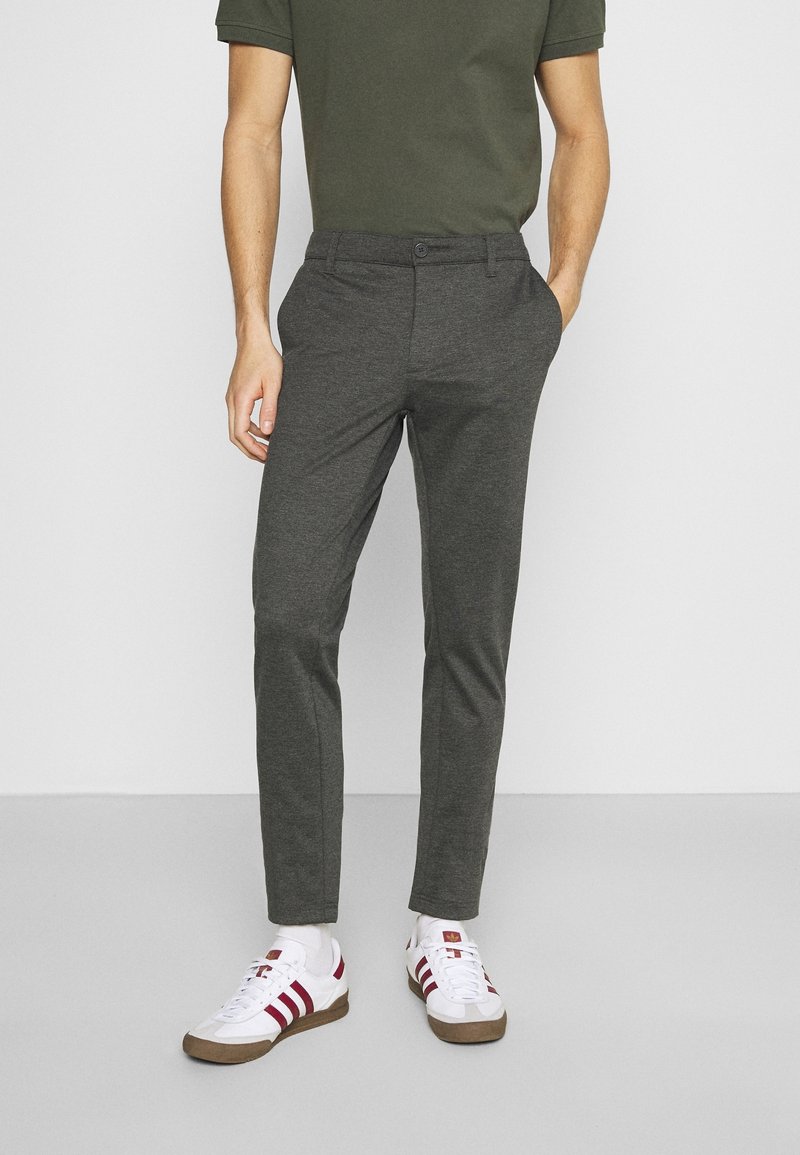 Pantalons gris tailleur avec une coupe slim, fabriqués à partir d'un tissu lisse, associés à une chemise verte et des baskets blanches avec des rayures rouges.