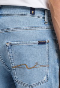 Jasnoniebieskie dżinsy z tylną kieszenią ozdobioną złotą haftą i małą granatową metką. Zniszczony detal w pobliżu krawędzi kieszeni.