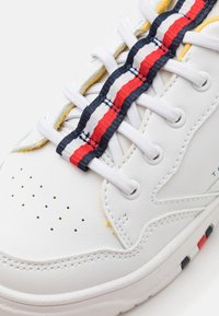 Sneaker bianca con tomaia in pelle testurizzata, lacci multicolori a strisce (rosso, bianco, blu scuro) e perforazioni per la traspirabilità.