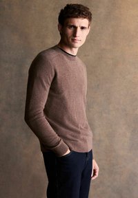 Brauner gestrickter Pullover mit langen Ärmeln und rundem Ausschnitt, kombiniert mit dunkelblauer Jeans. Dezente Textur und figurbetonte Design-Details.