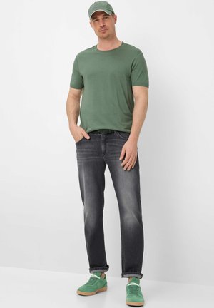 Mand iført grøn kasket, grøn t-shirt, sorte jeans og grønne sneakers, stående med den ene hånd i lommen foran en hvid baggrund.