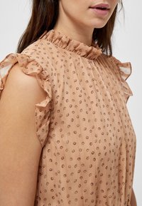 Hedvábná blůza v broskvové barvě s volánkovým vysokým výstřihem, krátkými volánkovými rukávy a texturovanou látkou s malými květinovými vzory po celé ploše.