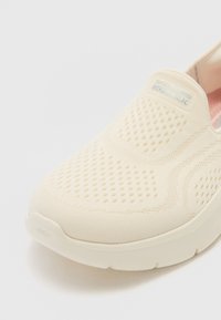 Adidași bej slip-on cu partea superioară din plasă, detalii texturate, talpă căptușită și eticheta "Skechers GOwalk" pe lateral.