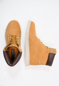 Botines marrones y beiges hechos de ante, con ojales dorados, suelas beige y un cuello acolchado en el tobillo. Acabado texturizado con colores contrastantes.