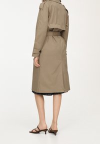 Femme portant un trench-coat beige ceinturé, une jupe noire mi-longue et des sandales marron à talons, vue de dos sur un fond blanc.