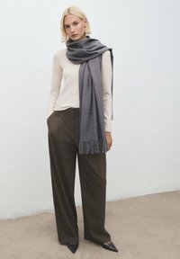 Sciarpa grigia appoggiata su una camicia color crema, abbinata a pantaloni marroni scuri. La sciarpa ha bordi frangiati e le scarpe sono nere e a punta. Texture lisce.