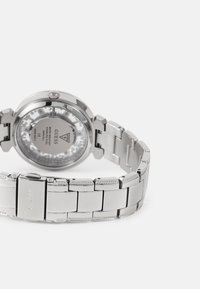 Hopea ruostumaton teräs kello harjatulla pinnalla, pyöreä runko, läpinäkyvä tausta ja linkkiketju, jossa on lukko ja koristeita.