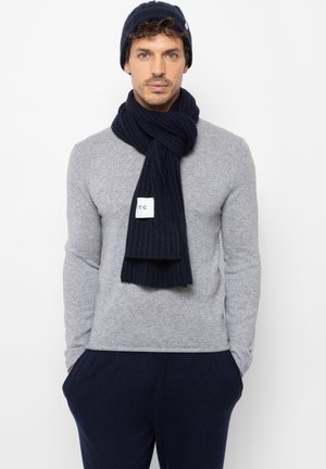 Marineblauwe geribbelde gebreide sjaal met een label, gedragen over een lichtgrijze trui. Het model heeft donker haar en draagt een bijpassende marineblauwe beanie.