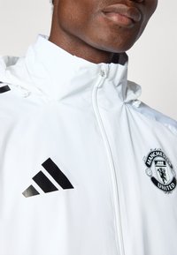 White waterproof jacket with a high collar, featuring a black Adidas logo and Manchester United crest on the left chest. Smooth texture.