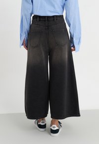 Calças de ganga pretas de perna larga com um efeito ombré gradual do escuro para o claro. Apresenta dois bolsos traseiros e é combinada com uma camisa de riscas azuis e ténis.