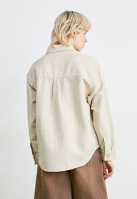 Light beige shirt, long sleeves, buttoned cuffs, back pleat detail, slightly curved hem, made of textured fabric.