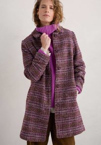 Cappotto a quadri marrone e rosa con una vestibilità strutturata, caratterizzato da grandi bottoni e un colletto, indossato sopra un maglione viola.