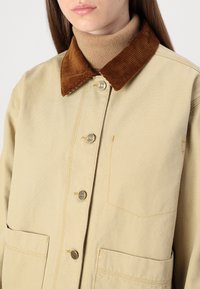 Giacca beige con colletto in velluto a coste marrone, bottoni metallici e due tasche frontali. Il tessuto ha una consistenza liscia e strutturata.