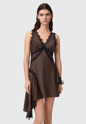 Jonge vrouw draagt een mouwloze bruine satijnen jurk met zwarte kanten afwerking en asymmetrische zoom, staand tegen een effen grijze achtergrond.