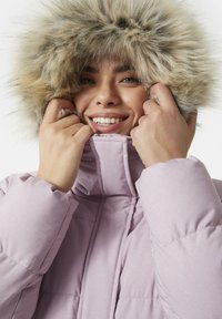 Svetle fialová péřová bunda s kapucí podšitou umělou kožešinou. Dlouhé rukávy a vysoký límec. Měkká textura s prošívaným vzorem. Usmívající se osoba.
