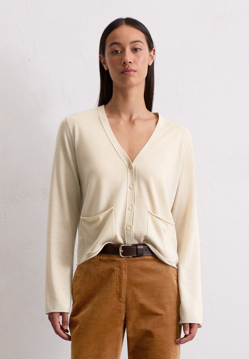 Cremfarbener Cardigan mit V-Ausschnitt, Knopfverschluss und zwei Fronttaschen. Weicher Stoff, lange Ärmel, über braunen Cordhosen getragen.