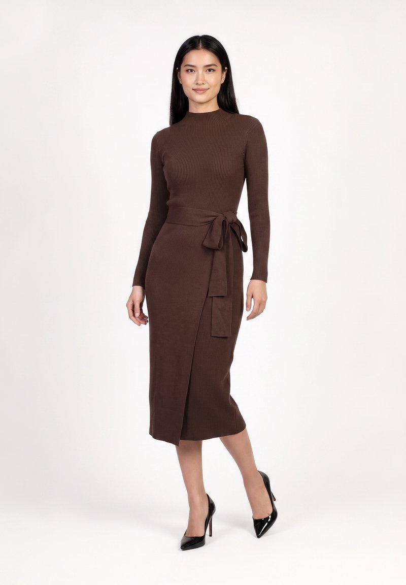 Femme portant une robe marron en tricot côtelé à manches longues avec une ceinture à la taille et des chaussures noires à talons hauts, debout devant un fond uni.