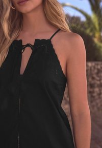 Haut noir sans manches avec bord en dentelle, lien ajustable au cou et un motif texturé le long de l'encolure. Tissu doux et coupe ample.