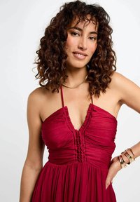 Vestito rosso senza maniche con texture a coste e scollatura profonda a V. Presenta tessuto arricciato sul seno e una gonna fluente. Bracciali dorati come accenti.