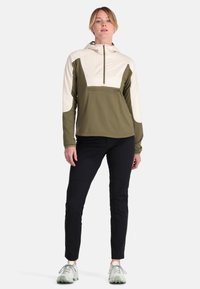 Leichter Kapuzenpullover in Olivgrün und Creme mit einer Fronttasche mit Reißverschluss. Kombiniert mit eng anliegenden schwarzen Hosen und strukturierten, hellen Schuhen.