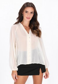 Blusa transparente color beige claro con cierre de botones en el frente, mangas largas y escote fruncido; combinada con una falda negra que presenta una abertura lateral.