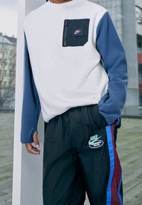 Vit och blå långärmad sweatshirt med frontfickor med dragkedja; ihop med svarta byxor med färgade ränder och Nike-logotyp.