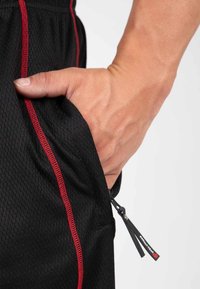 Zwarte sportshorts van een gestructureerde stof, voorzien van rode stiksels en een ritszak. Hand geplaatst in de zakdetail.