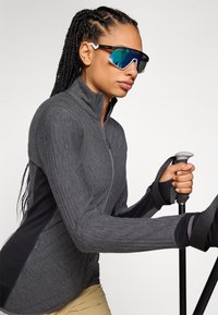 Dark grey, long-sleeve, zip-up jacket with textured fabric, featuring a black panel. Model holds a trekking pole, wearing mirrored sunglasses.