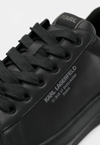 Zapatillas de cuero negro con cordones redondeados, parte superior texturizada y detalles del logo en relieve en el costado, junto con elementos de diseño minimalista.