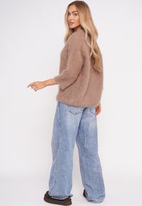 Eine Frau mit langen blonden Haaren, die einen weichen braunen Pullover, lockere hellblaue Jeans und dunkle braune Schuhe trägt, steht vor einem weißen Hintergrund.