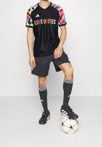 Černý sportovní dres s barevnými vzorovanými rukávy, s nápisem "LÁSKA SPOJUJE" vytištěným tučnými písmeny. V kombinaci se šedými šortkami, černými ponožkami a kopačkami, postaven na fotbalovém míči.