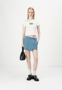 Camiseta blanca corta con gráfica de frutas verdes, falda envolvente de mezclilla azul claro, bolso negro, calcetas blancas y calzado negro. Estilo simple y casual.