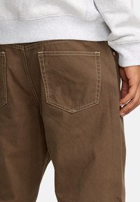 Bruine katoenen broek met een gestructureerde afwerking, vijfzakkenontwerp en contrasterende stiksels. G gedragen met een grijze sweatshirt.