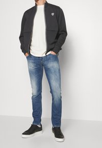 Námornická zipová bunda s bílými akcenty, bílý tričko, světlé modré džíny a černé tenisky. Slim-fit silueta a pohodlný design.