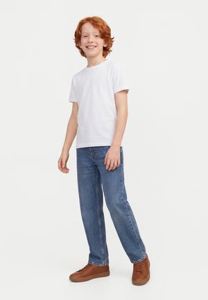 Rothaariger Junge mit weißem T-Shirt, blauer Jeans und braunen Turnschuhen, der vor einem einfachen weißen Hintergrund steht und lächelt.
