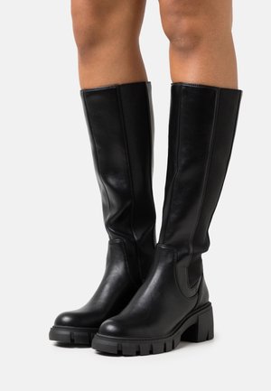 Bottes en cuir noires montant jusqu'aux genoux avec semelles crantées épaisses portées sur des jambes nues, sur un fond blanc uni.