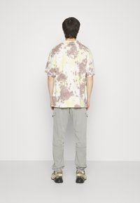 Tricou cu mânecă scurtă, tie-dye în nuanțe de galben pastel și maro, asortat cu pantaloni cargo gri și adidași negri/verzi. Fundal simplu.