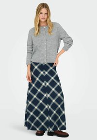 Cárdigan gris de punto con cuello redondo y botones, combinado con una falda larga de cuadros azul marino con líneas blancas y azules claro. Botas gruesas marrones.