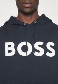 Muž oblečený v tmavě námořnické mikině s bílým logem BOSS umístěným na středu hrudi a s tkaničkami visícími z kapuce.