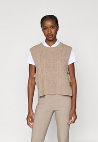 Pulover bej fără mâneci, tricotat, cu model cu cabluri și tiv striat, purtat peste o cămașă albă cu guler. Modelul are părul împletit.