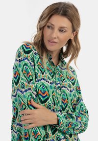 Raštuotas mygtukų marškiniai, pasižymintys ryškiomis žaliomis, mėlynomis, oranžinėmis ir baltomis spalvomis, su tekstūruota medžiaga ir apykakle.