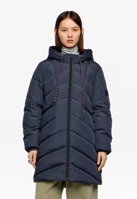 Kobieta w granatowej, pikowanej kurtce z kapturem, zamkiem b�łyskawicznym i ściągaczami, w jasnooliwkowych spodniach, stojąca na białym tle.