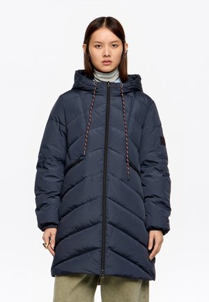 Kobieta w granatowej, pikowanej kurtce z kapturem, zamkiem błyskawicznym i ściągaczami, w jasnooliwkowych spodniach, stojąca na białym tle.