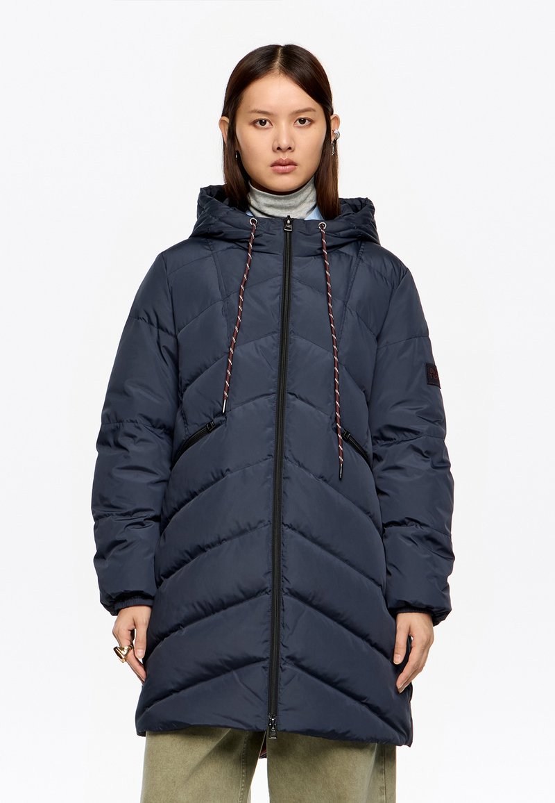 Kobieta w granatowej, pikowanej kurtce z kapturem, zamkiem błyskawicznym i ściągaczami, w jasnooliwkowych spodniach, stojąca na białym tle.