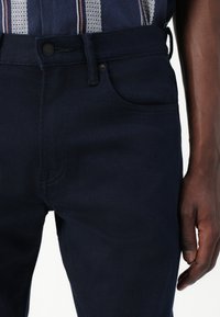 Jeans din denim bleumarin cu o textură netedă, având o buzunar frontal, bucle pentru curea și închidere cu nasture. Detalii subtile de coasere vizibile.